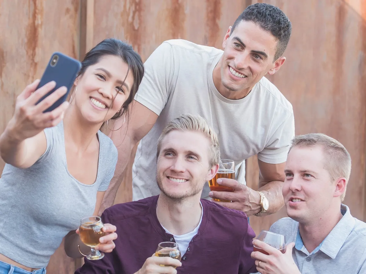 Vier vrienden heffen hun biertjes voor een selfie op de binnenplaats van een brouwerij, onder de lichtslingers